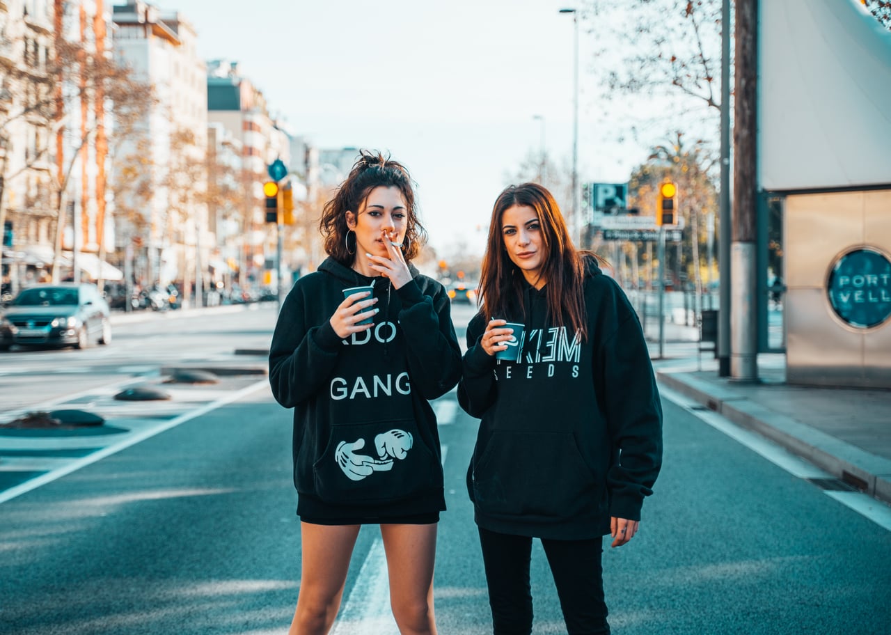 Dues dones joves amb dessuadores streetwear dempeus al mig d'una avinguda de Barcelona, una fumant amb got de cartró, retrat urbà editorial.