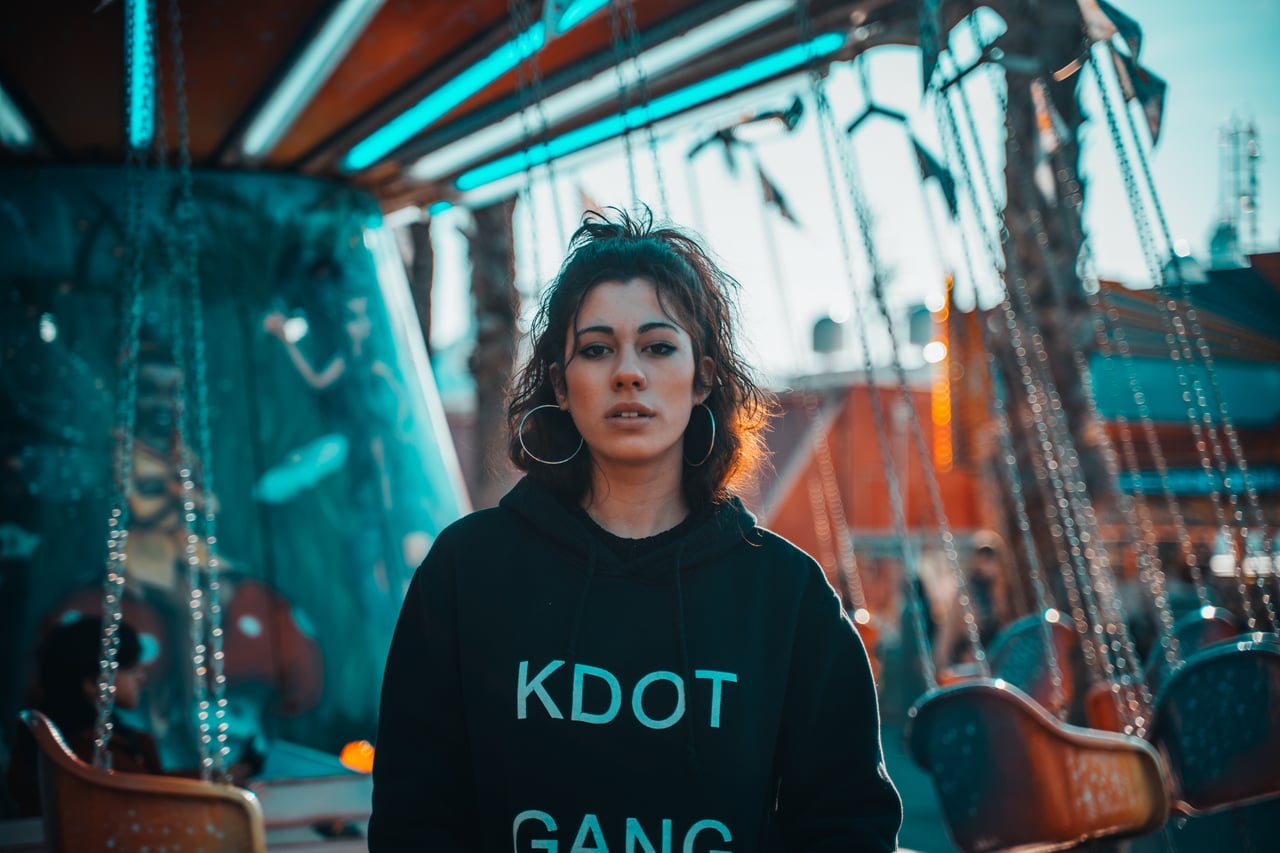 Dona jove amb dessuadora negra streetwear en una fira, mirada directa, atraccions cromàtiques taronja i turquesa desenfocades al fons.