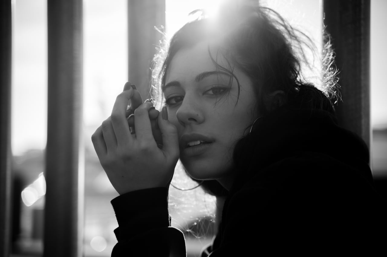 Dona jove amb dessuadora fosca recolzant les mans a la cara amb expressió pensativa, contrallum fort i llums bokeh, blanc i negre.