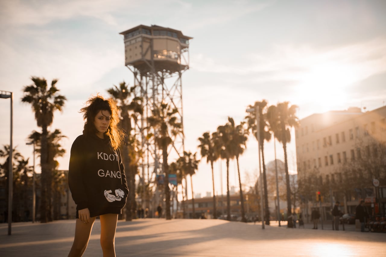 Dona jove amb dessuadora negra streetwear caminant per una plaça de Barcelona al capvespre, torre Sant Sebastià del telefèric i palmeres de fons.