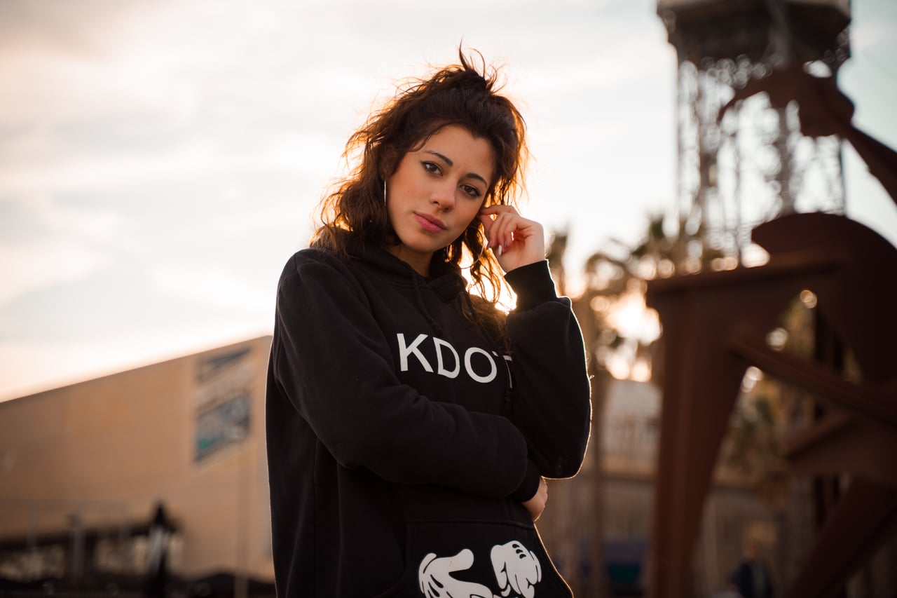 Dona jove amb dessuadora negra Kdot Gang i mà recolzada al pols, mirada confiada a càmera, exterior urbà de Barcelona al capvespre.