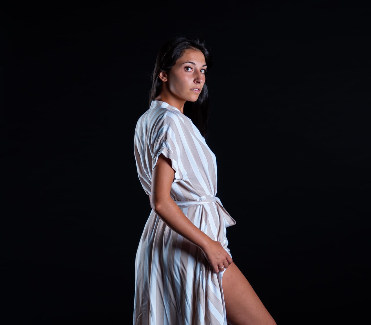 Dona jove amb vestit camiser a ratlles blanques i beix de perfil amb cama sortint, fons d'estudi negre low-key.