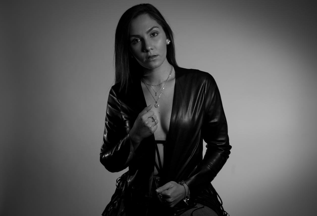 Dona jove amb jaqueta de cuir negra oberta sobre tors nu cobert només per collarets i mans, retrat en blanc i negre sobre fons d'estudi gris.