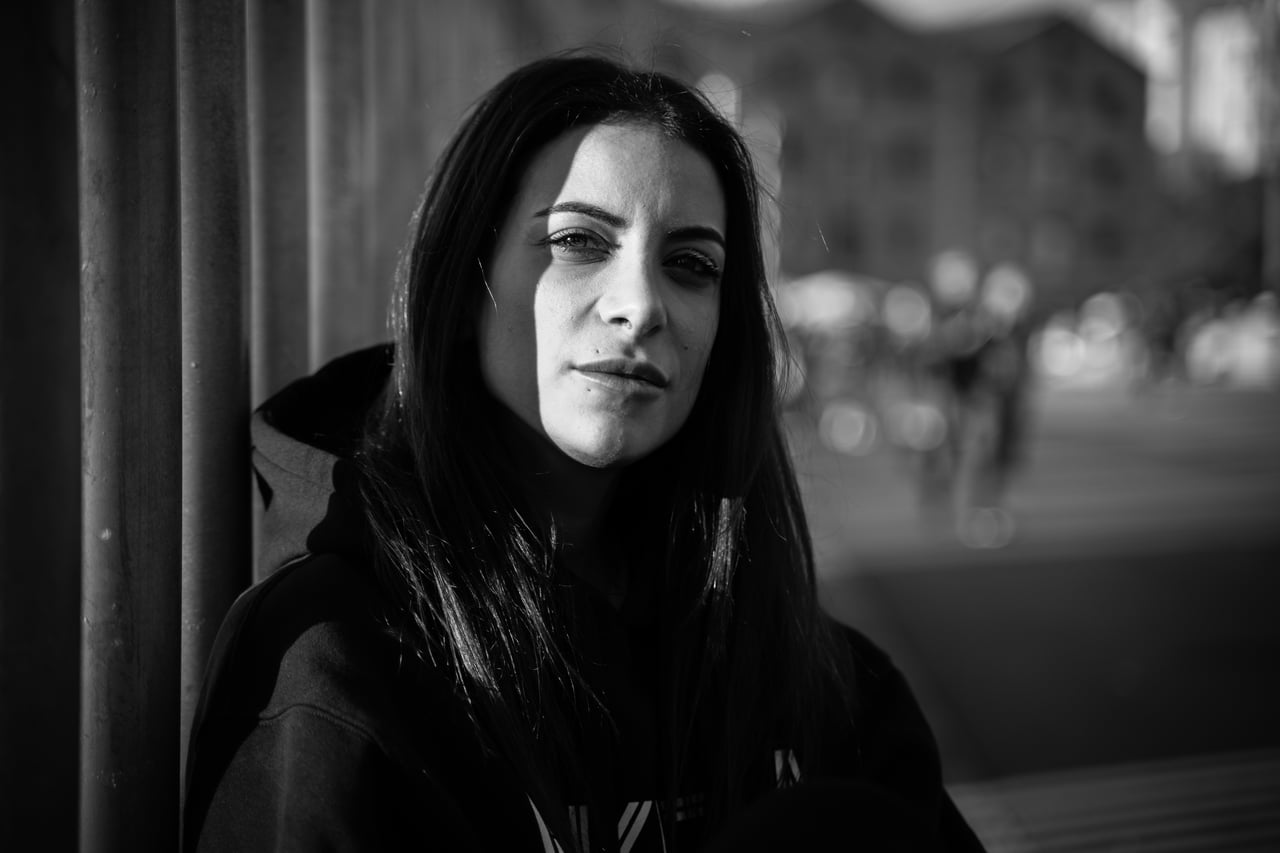 Dona jove amb dessuadora fosca recolzada contra una columna metàl·lica, mirada directa a càmera, carrer urbà desenfocat al fons, retrat en blanc i negre.