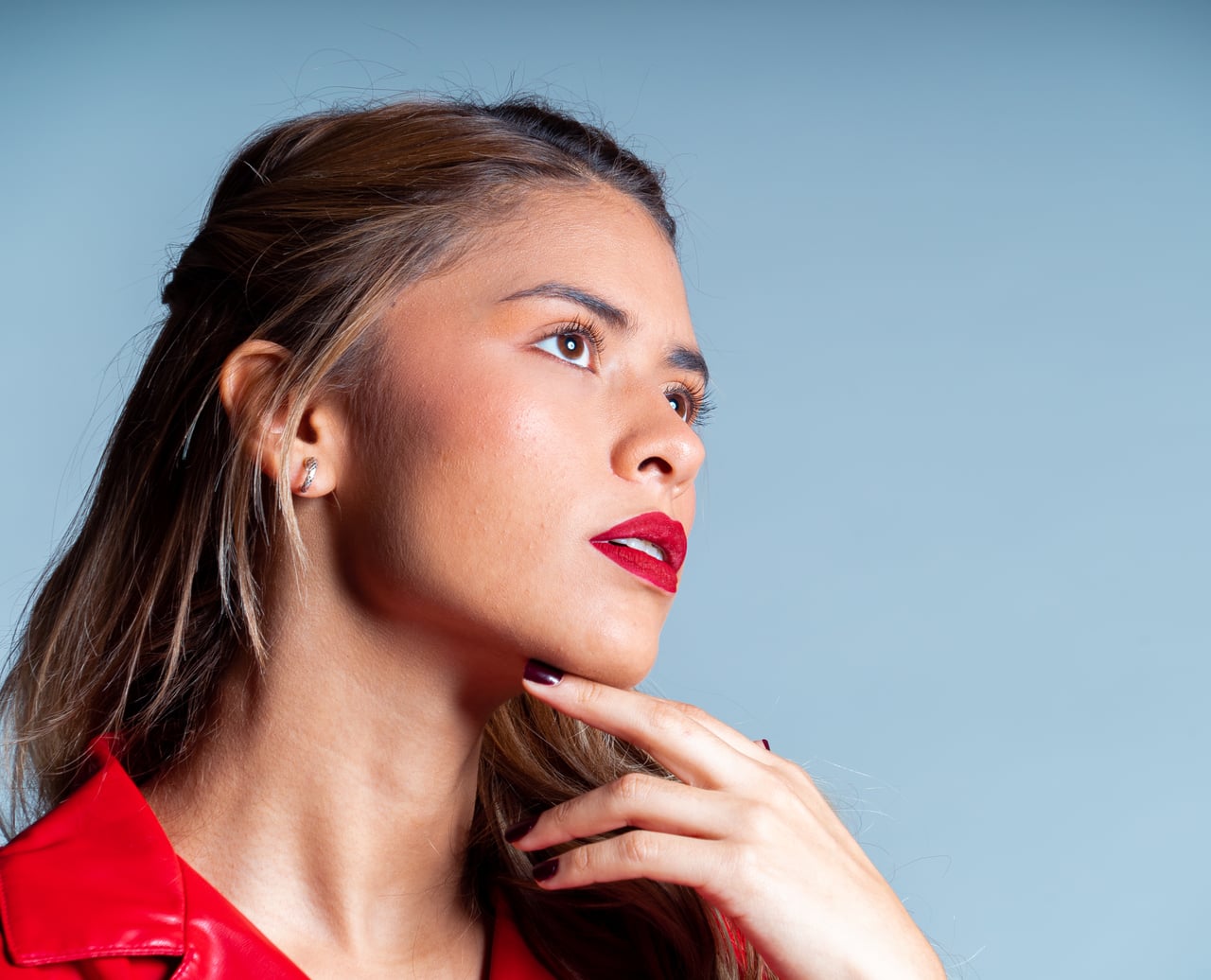 La Bianca de perfil mirant cap amunt amb la mà recolzada a la barbeta, cabellera rossa, peça de vinil vermell i llavis vermells, sobre fons d'estudi blau clar.