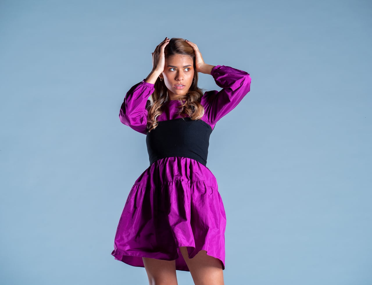 La Bianca Díaz es porta totes dues mans al cap amb un gest expressiu, enquadrament de mig cos, lluint un vestit curt fúcsia de mànigues bufades amb banda ampla negra cenyida a la cintura, sobre fons d'estudi blau.