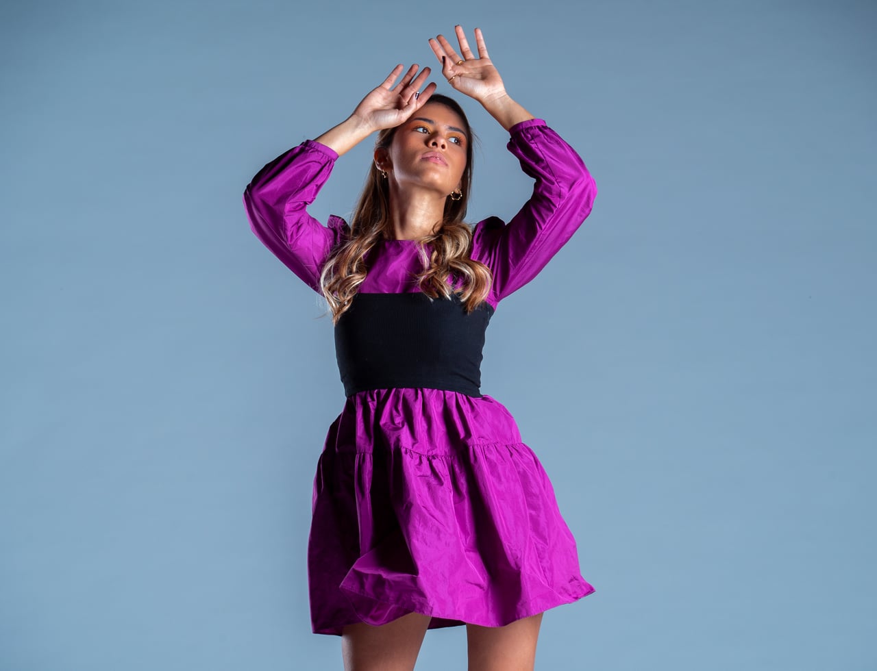 La Bianca Díaz aixeca tots dos braços per damunt del cap en una pose editorial dinàmica, enquadrament de mig cos, amb un vestit curt fúcsia de mànigues bufades i banda ampla negra cenyida a la cintura, sobre fons d'estudi blau.