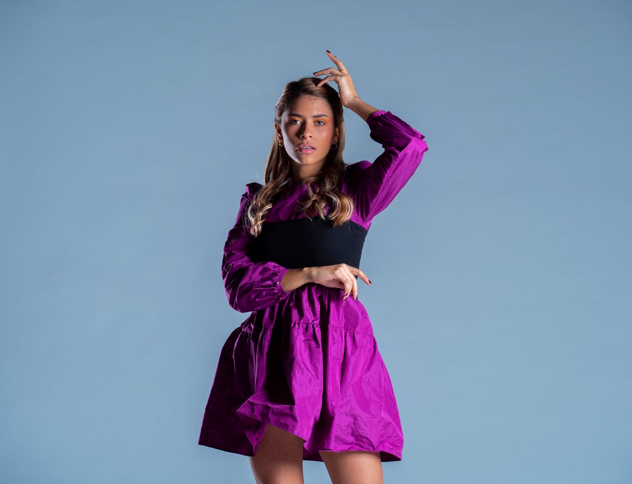 La Bianca Díaz posa amb una mà a prop del cap i l'altra fregant la cintura, enquadrament de mig cos, vestint un vestit curt fúcsia de mànigues llargues bufades amb banda ampla negra cenyida a la cintura, sobre fons d'estudi blau.