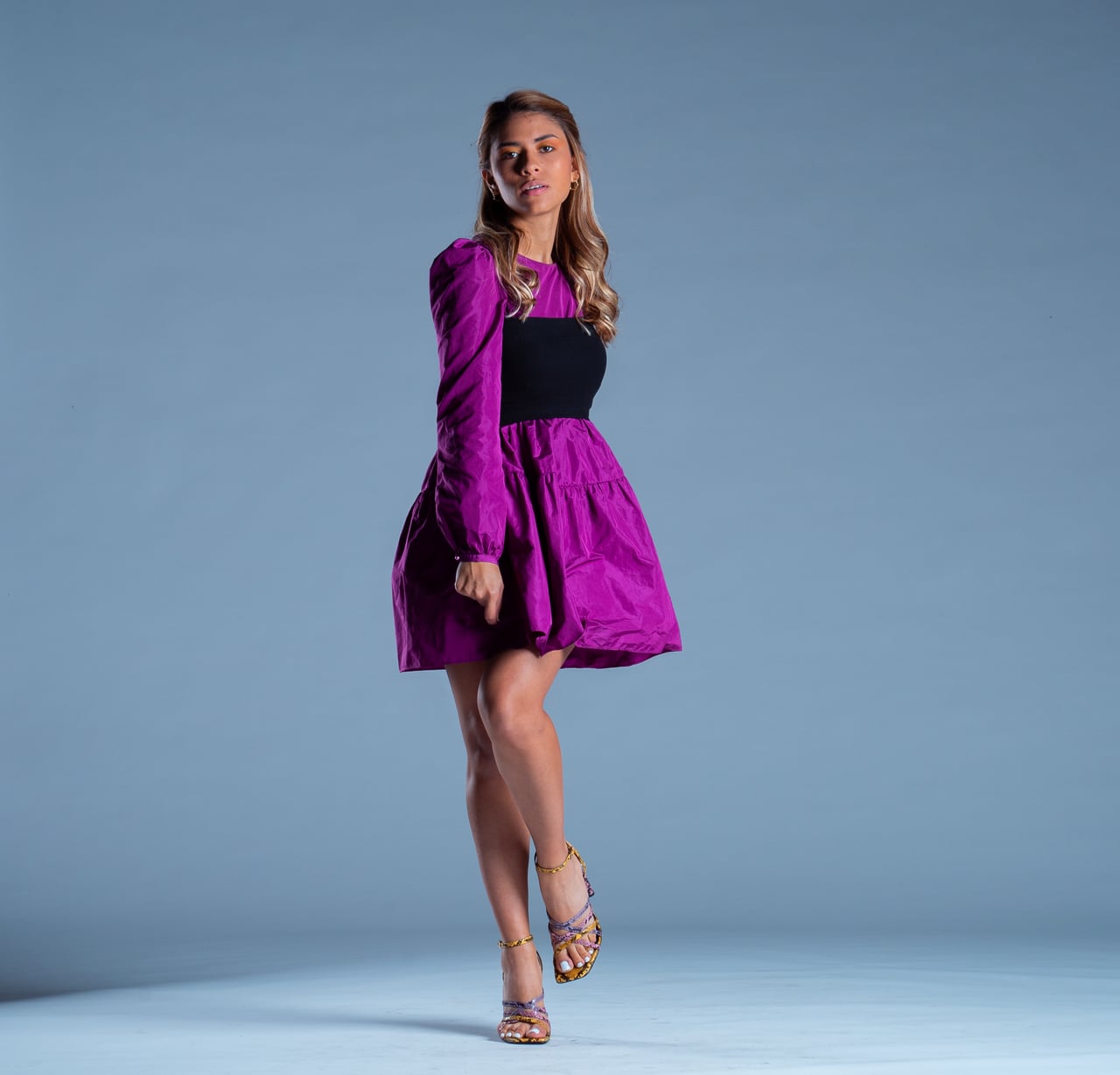 La Bianca Díaz avança cap a la càmera amb una cama flexionada en una pose editorial de cos sencer, amb vestit curt fúcsia de mànigues bufades, banda ampla negra a la cintura i sandàlies de taló estampades, sobre fons d'estudi blau.