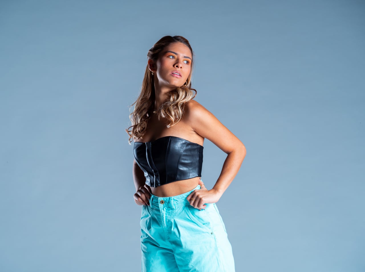 La Bianca Díaz posa de mig cos amb les mans als malucs i la mirada de costat, vestida amb un top cotilla escotat de pell negra i un pantaló ample de cintura alta en turquesa, sobre un fons d'estudi blau acer.