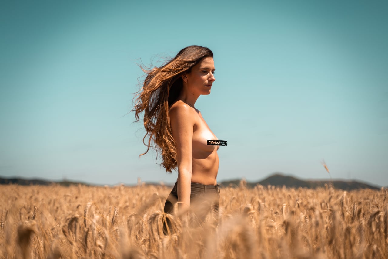 Dona en topless de perfil caminant per un camp de blat daurat amb el cabell volant al vent, censura gràfica sobre el mugró visible.