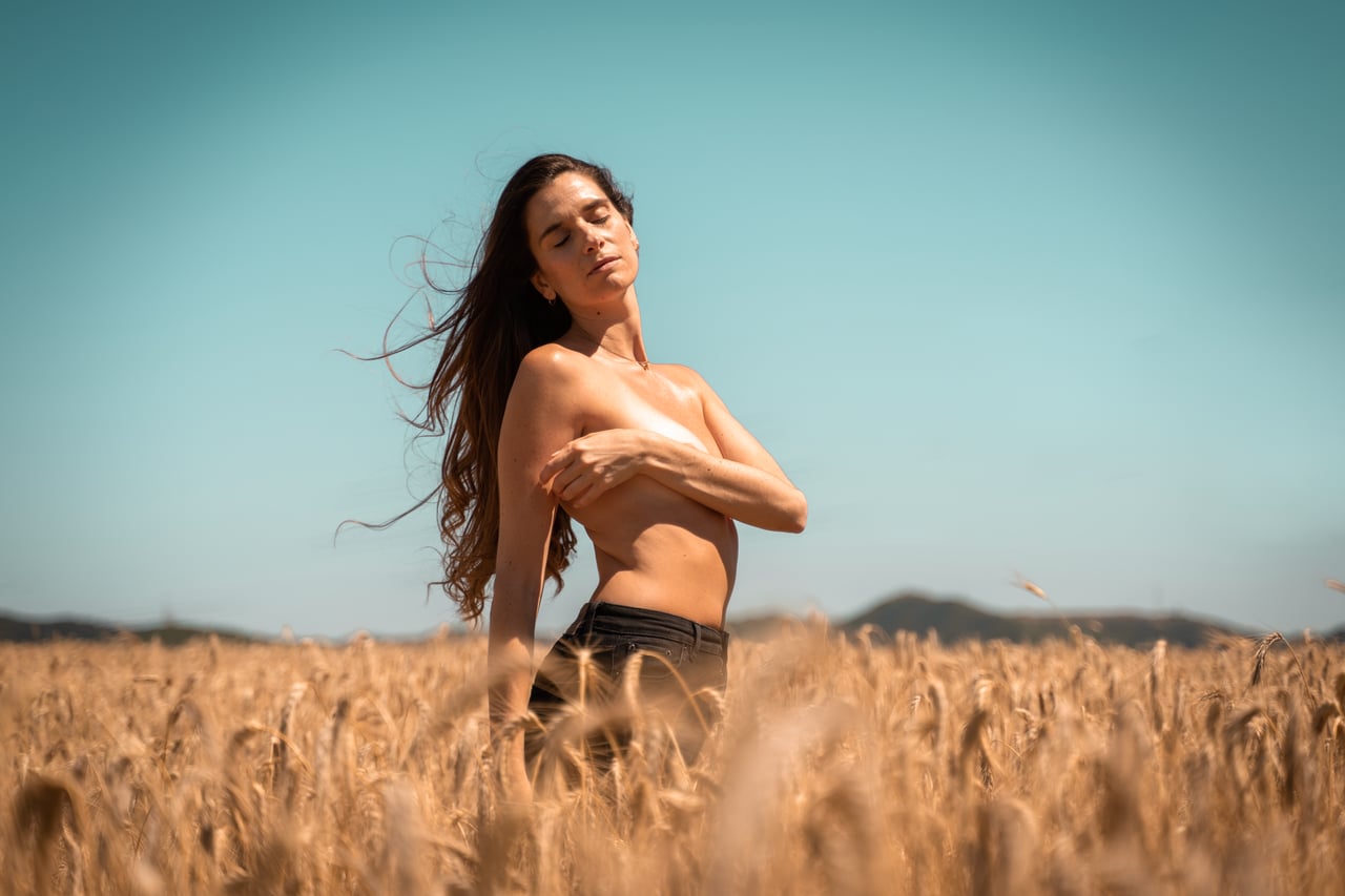 Dona en topless en un camp de blat daurat cobrint-se el pit amb un braç creuat, ulls tancats i cabell al vent, muntanyes al fons.
