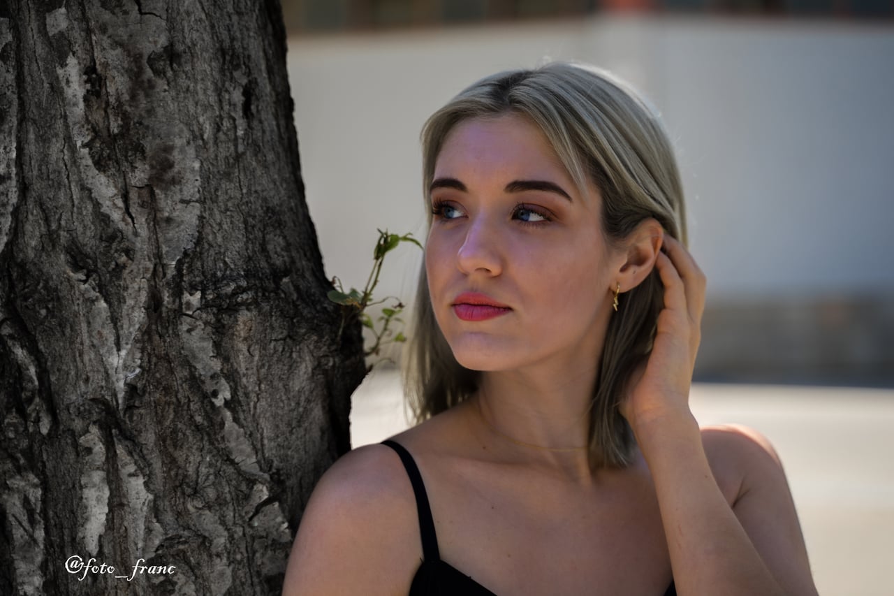 Dona jove rossa amb ulls blaus i top negre de tirants, mirant de costat amb la mà recolzada al cabell, al costat d'un tronc d'arbre en exterior amb llum natural càlida.