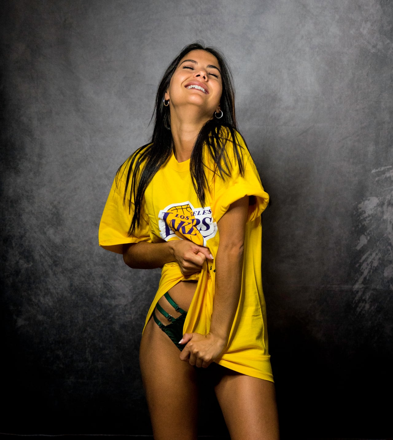 Dona amb samarreta groga dels Los Angeles Lakers rient amb el cap tirat enrere, fons d'estudi gris texturitzat.