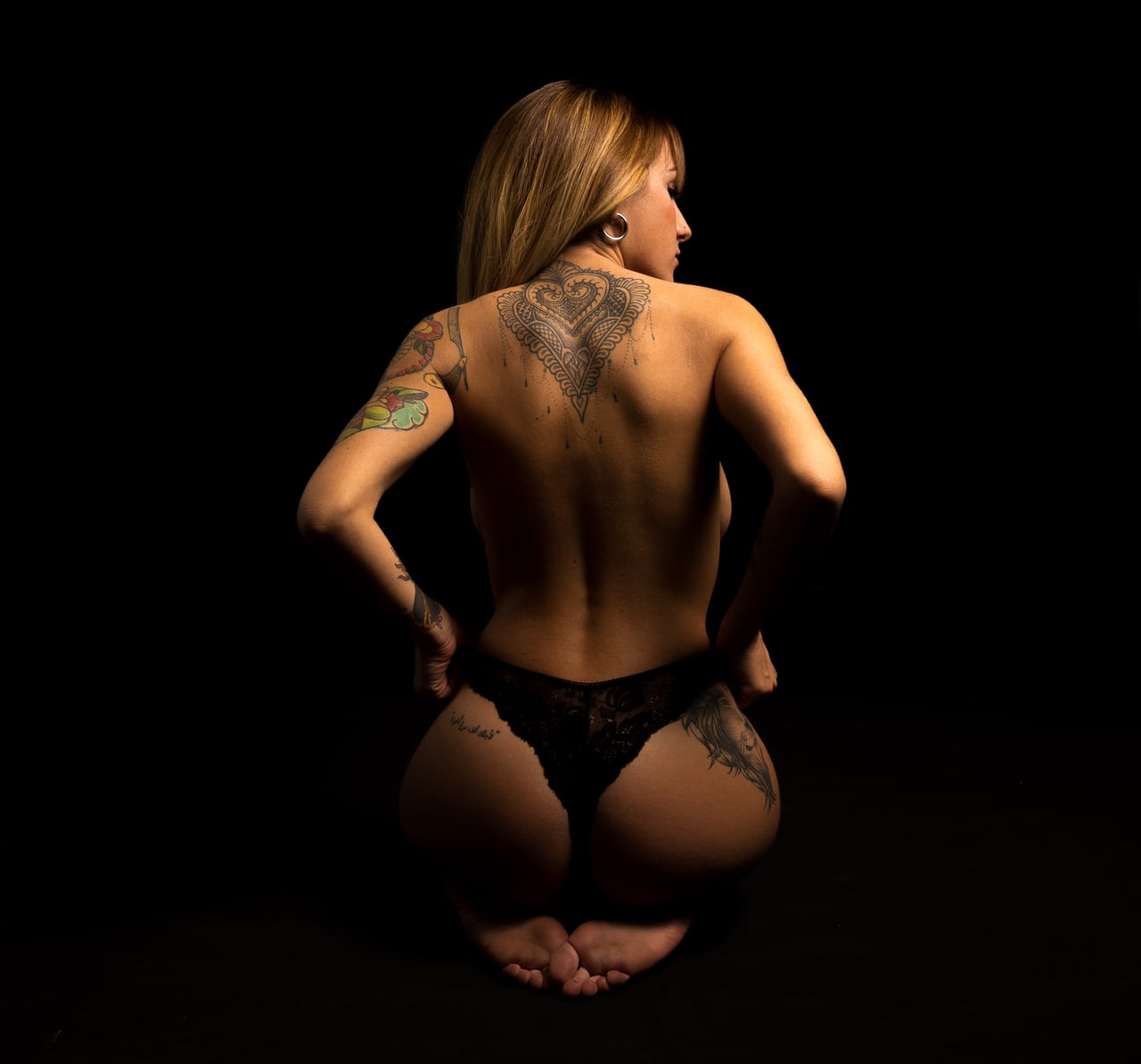 Dona agenollada d'esquena en topless amb calça de puntes negra, mostrant un tatuatge ornamental en forma de cor mandala a la zona alta de l'esquena, fons d'estudi completament negre.