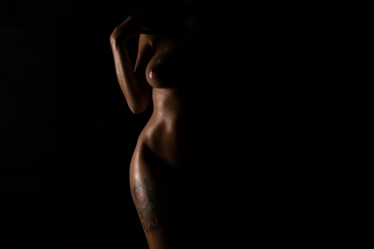 Silueta lateral de cos femení nu il·luminat de costat, braç elevat sobre el cap i tatuatge floral colorit a la cama, fons d'estudi completament negre.