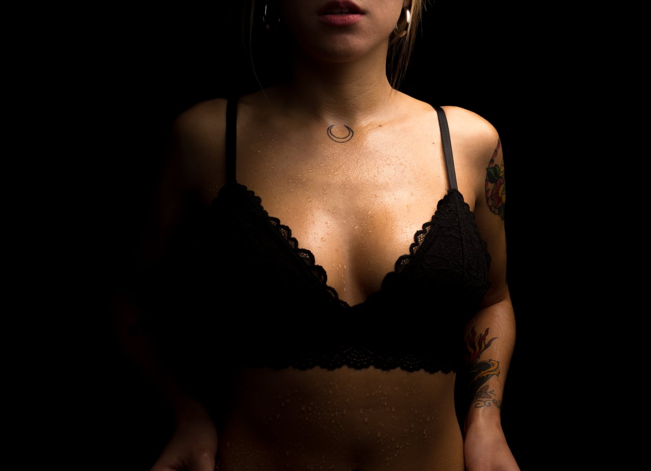 Detall de mig cos de dona rossa amb sostenidor negre de puntes, gotes d'aigua relliscant per l'escot i ventre, tatuatges colorits a espatlla i braç, fons d'estudi negre.