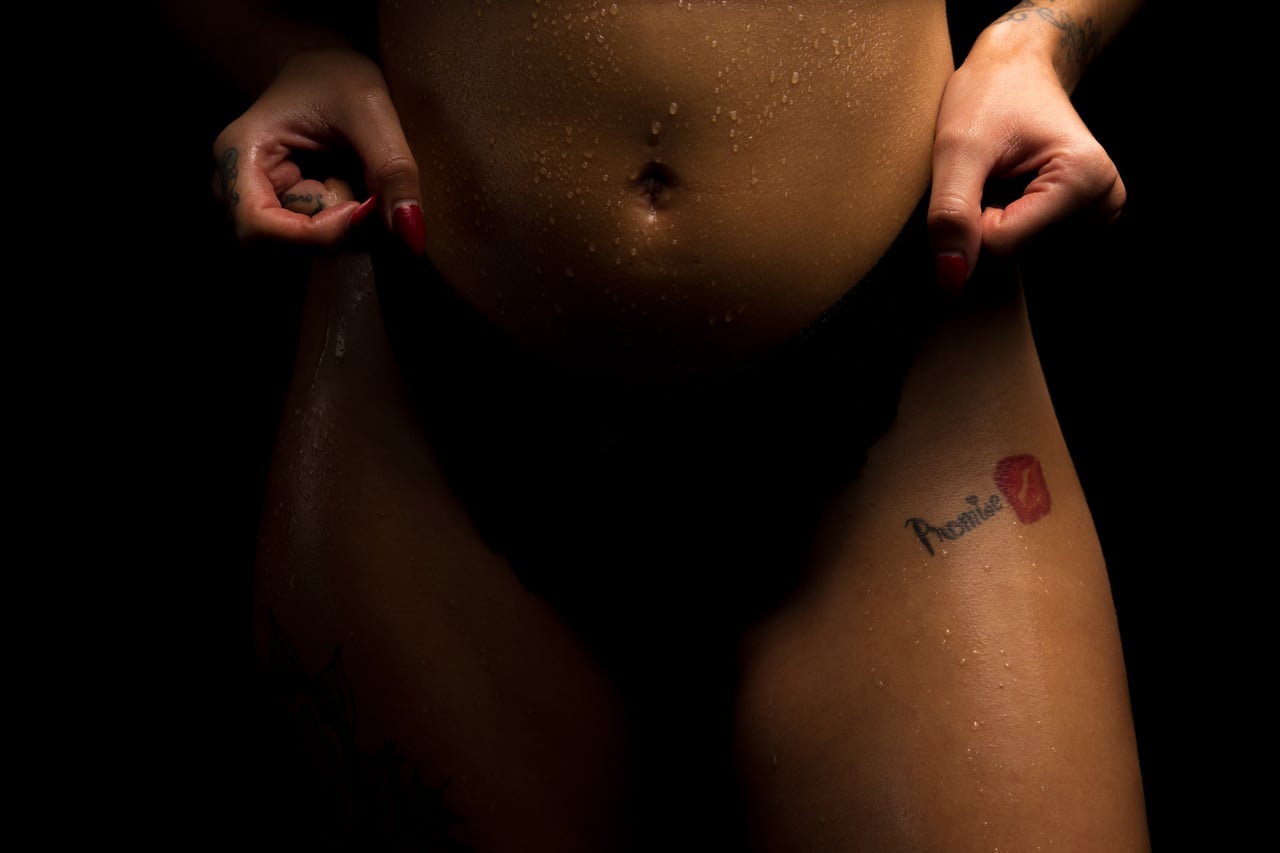 Detall del ventre femení amb calça negra i mans als malucs, gotes d'aigua sobre la pell, tatuatge 'Promise' i petjada de llavis vermella a la cuixa, fons d'estudi negre.