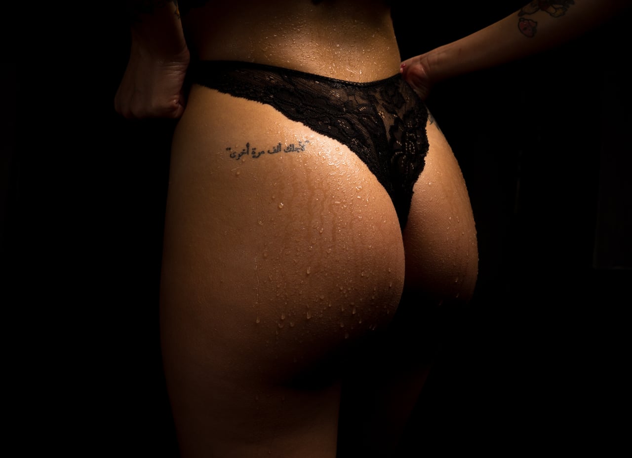 Detall de glutis femenins amb calça negra de puntes, gotes d'aigua sobre la pell i tatuatge en cal·ligrafia àrab a l'anca, fons d'estudi negre.
