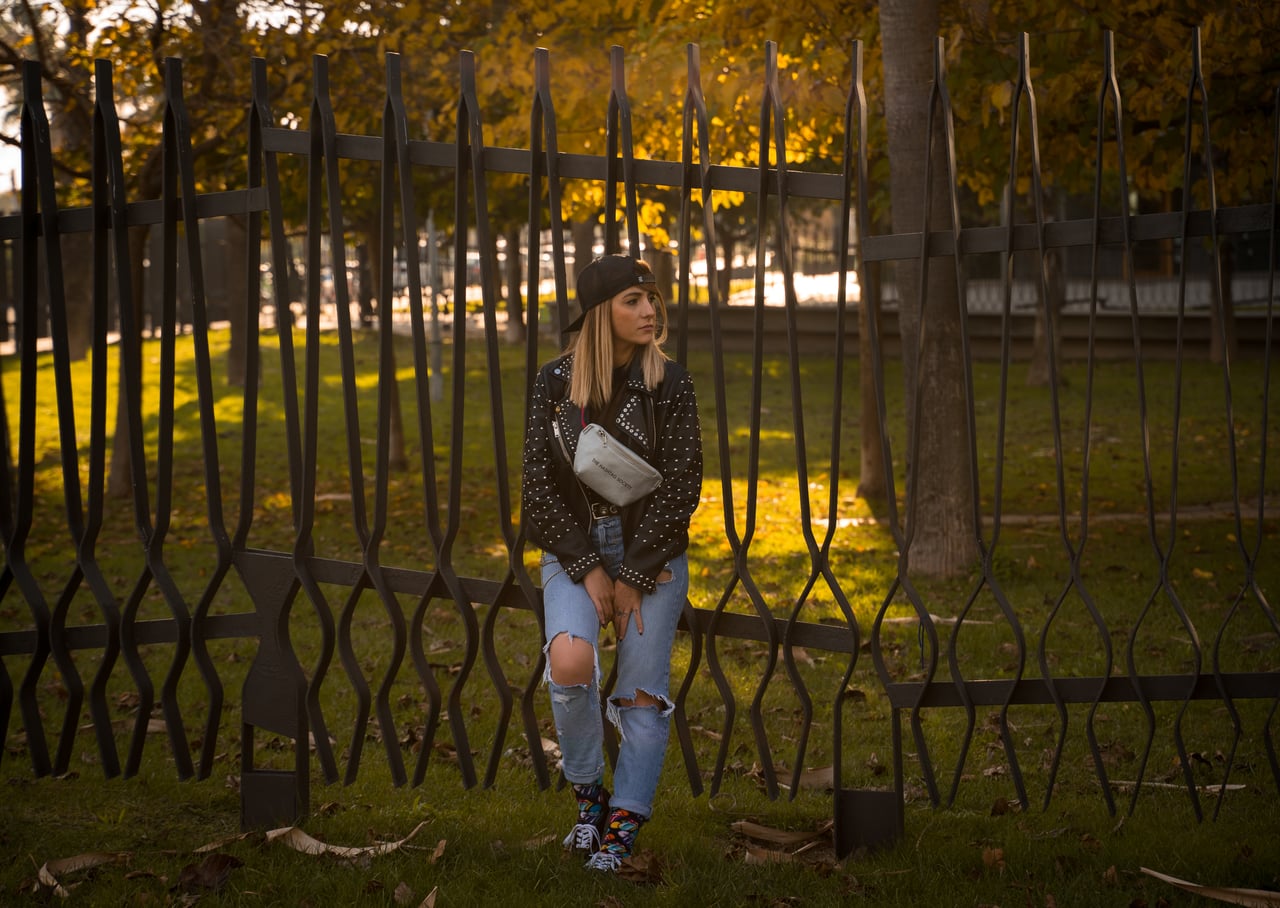 Dona rossa recolzada en una tanca negra forjada amb gorra negra, jaqueta de cuir amb tatxes, texans esquinçats i ronyonera blanca, parc urbà tardorenc amb arbres daurats de fons.