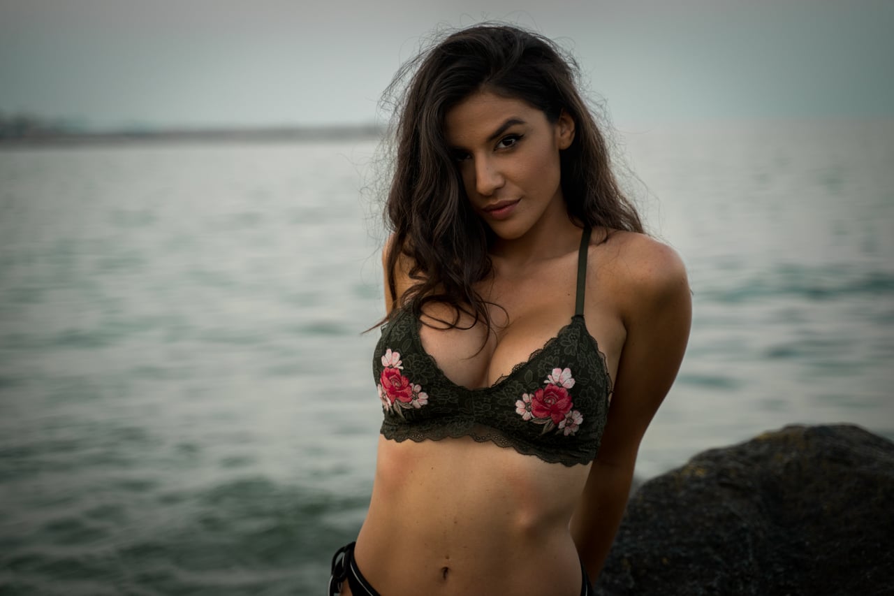 Dona amb bikini de puntes verd amb flors rosades brodades, al costat del mar gris en dia ennuvolat, mirada directa a càmera.