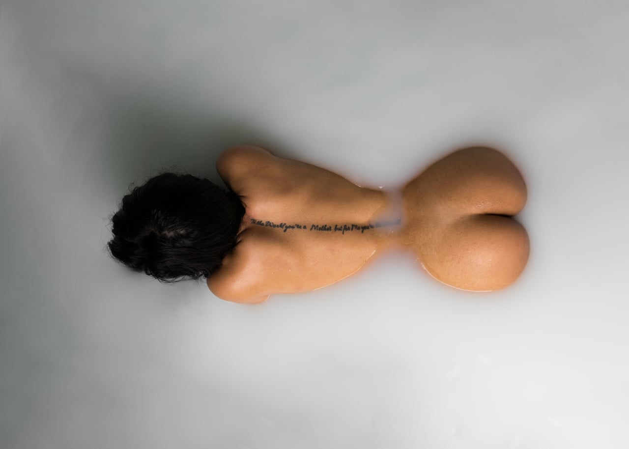 Vista zenital de dona en banyera plena de llet, d'esquena, amb un tatuatge de text en cursiva a l'esquena i glutis visibles emergint de la superfície.
