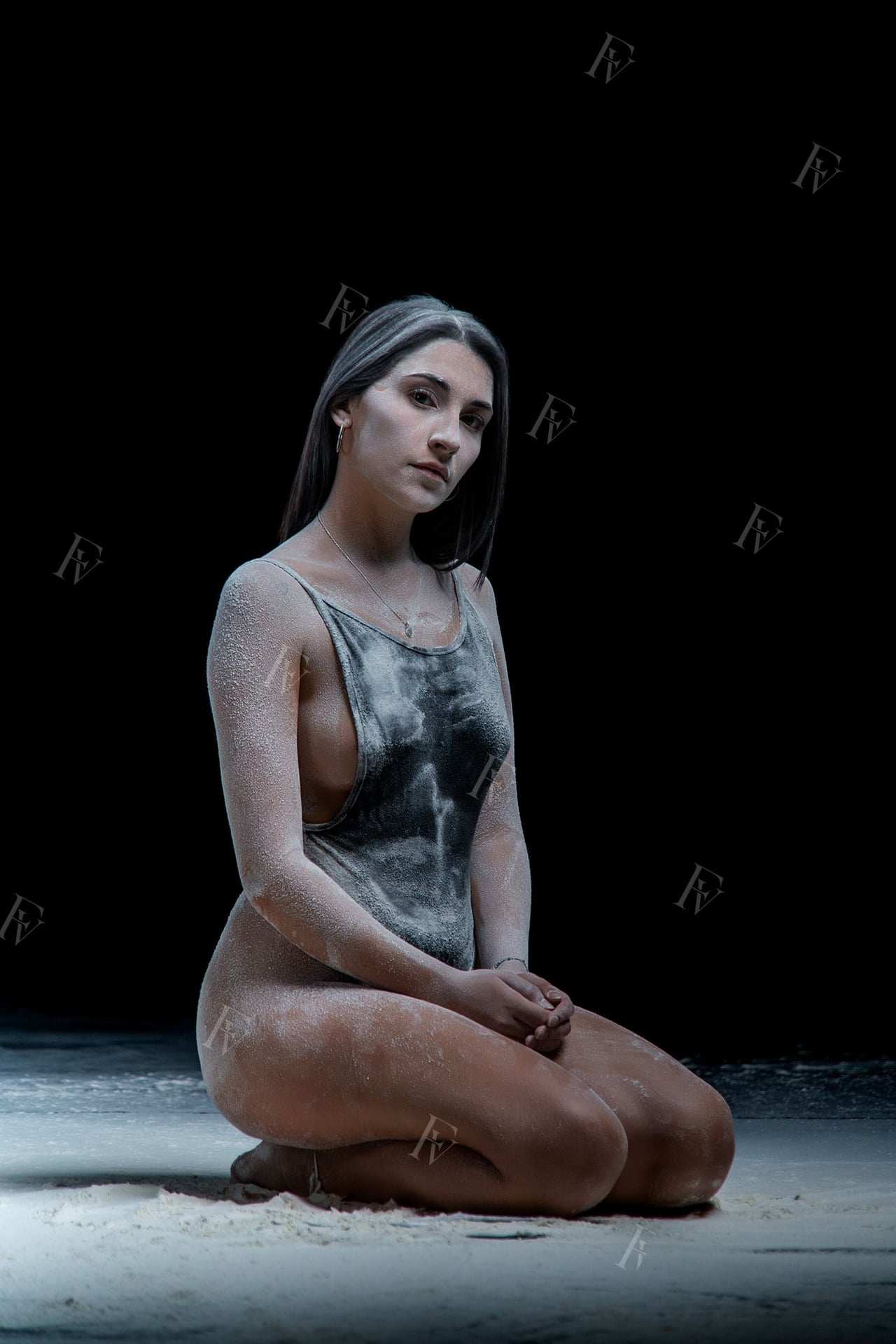 Ballarina amb body en pose editorial amb farina blanca envoltant-la, fons d'estudi negre low-key.