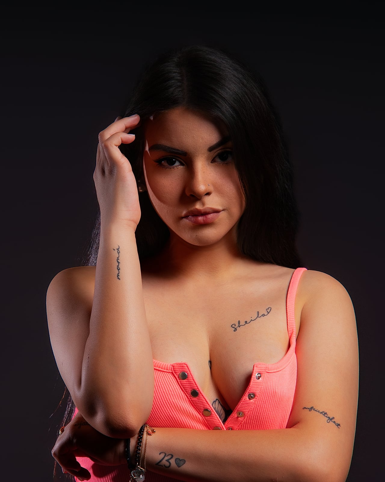 Dona jove amb top neó rosa, mà recolzada al cap, mirada directa, fons d'estudi negre low-key.