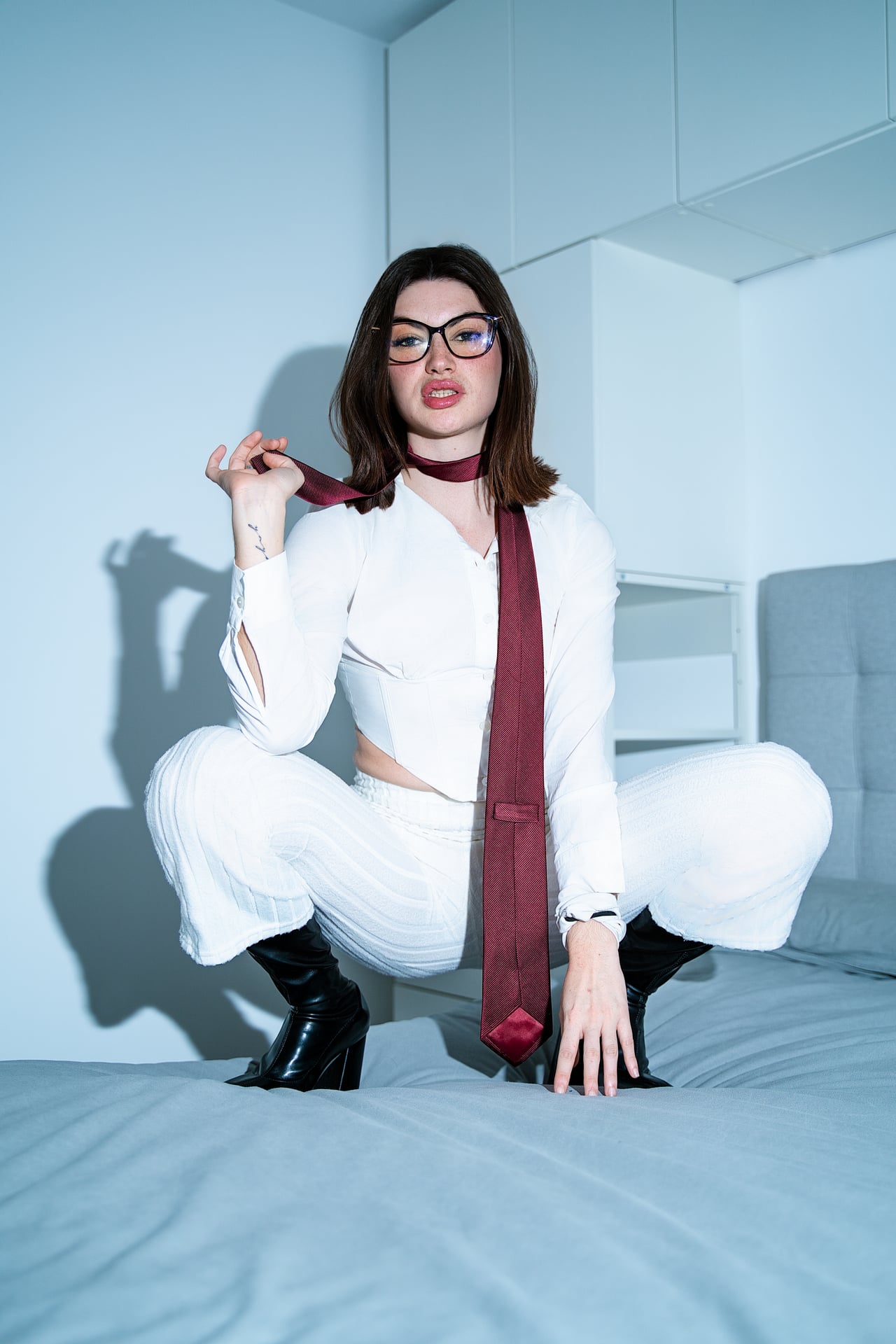 Dona jove amb conjunt blanc (camisa curta i pantaló ample), corbata fina bordeus i botes negres, agenollada sobre llit gris en dormitori lluminós.