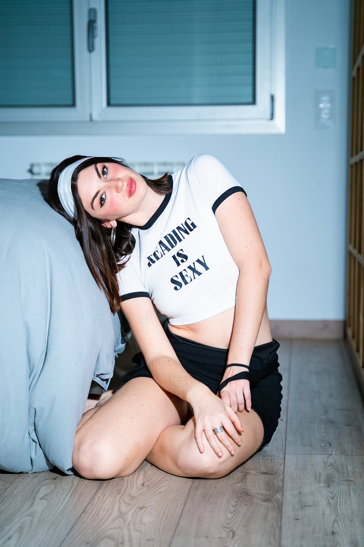 Dona jove amb diadema blanca i samarreta "Reading is sexy" recolzada contra un llit gris en dormitori lluminós.