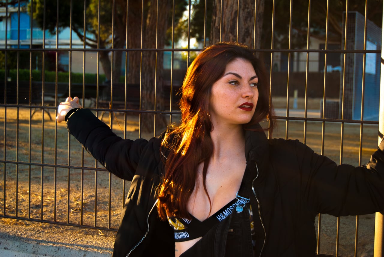 Dona jove pèl-roja amb cabell llarg, llavis vermells, bralette estampada H&M Moschino i bomber negra oberta recolzada contra una reixa metàl·lica d'un parc al capvespre, contrallum daurat entre els pins.