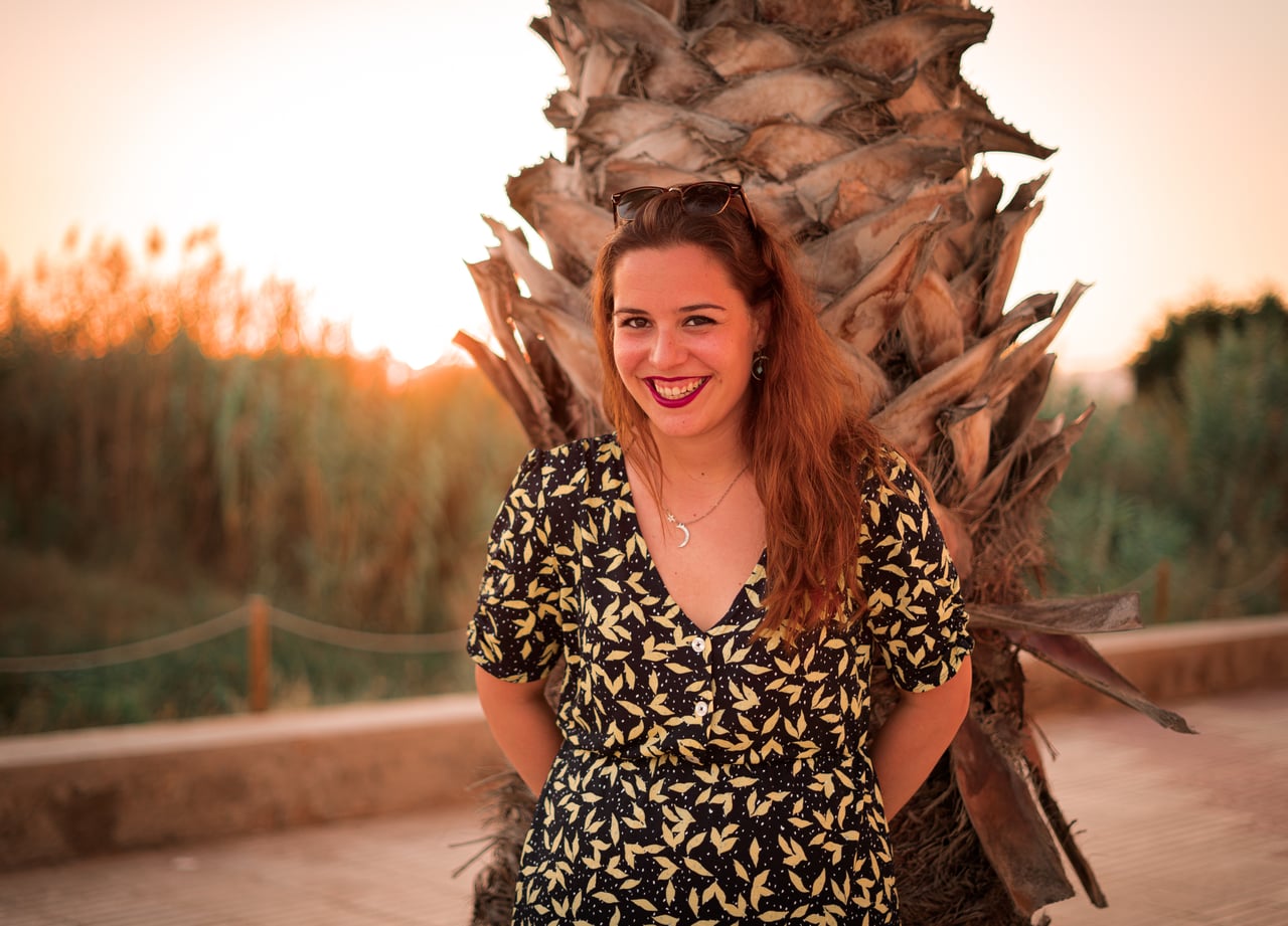 Retrat de mig cos amb vestit negre de fulles grogues i ulleres de sol al cap, somrient de cara amb una palmera i canyes il·luminades pel sol al fons.