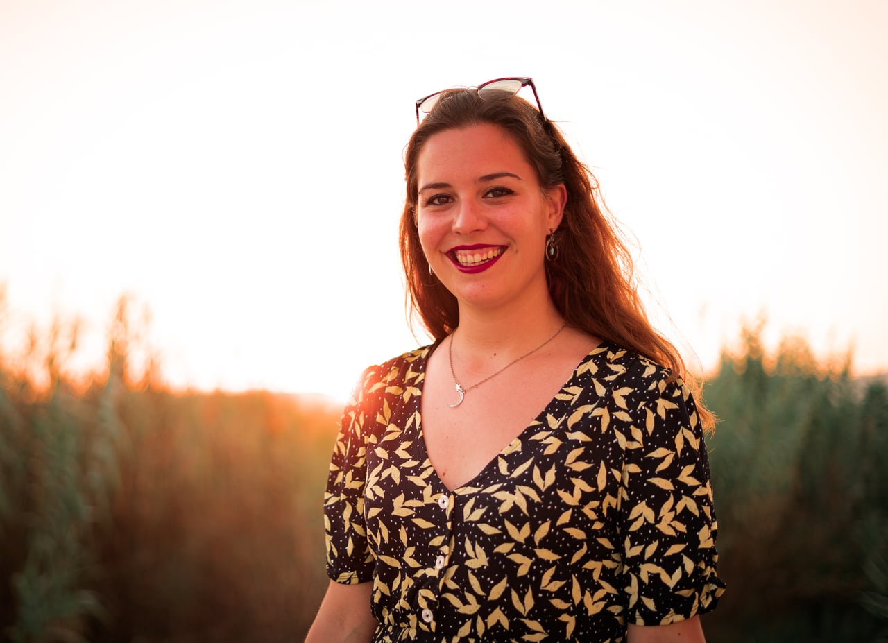 Retrat de mig cos somrient a càmera, vestit negre amb estampat de fulles grogues, ulleres de sol al cap i collaret de lluna, davant un fons de canyes al capvespre.