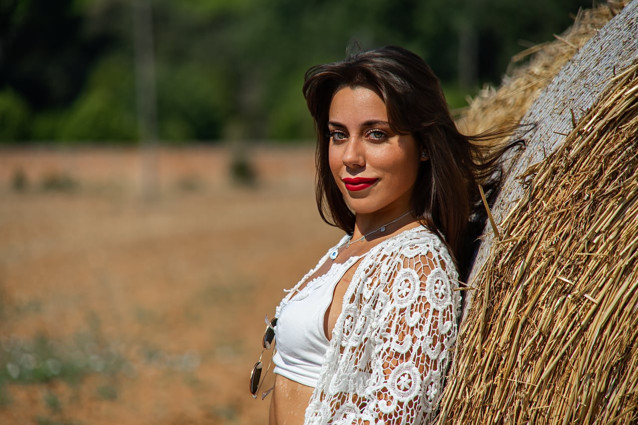 Odei8 mira a càmera al costat d'una bala de palla, amb top blanc i fons rural desenfocat.