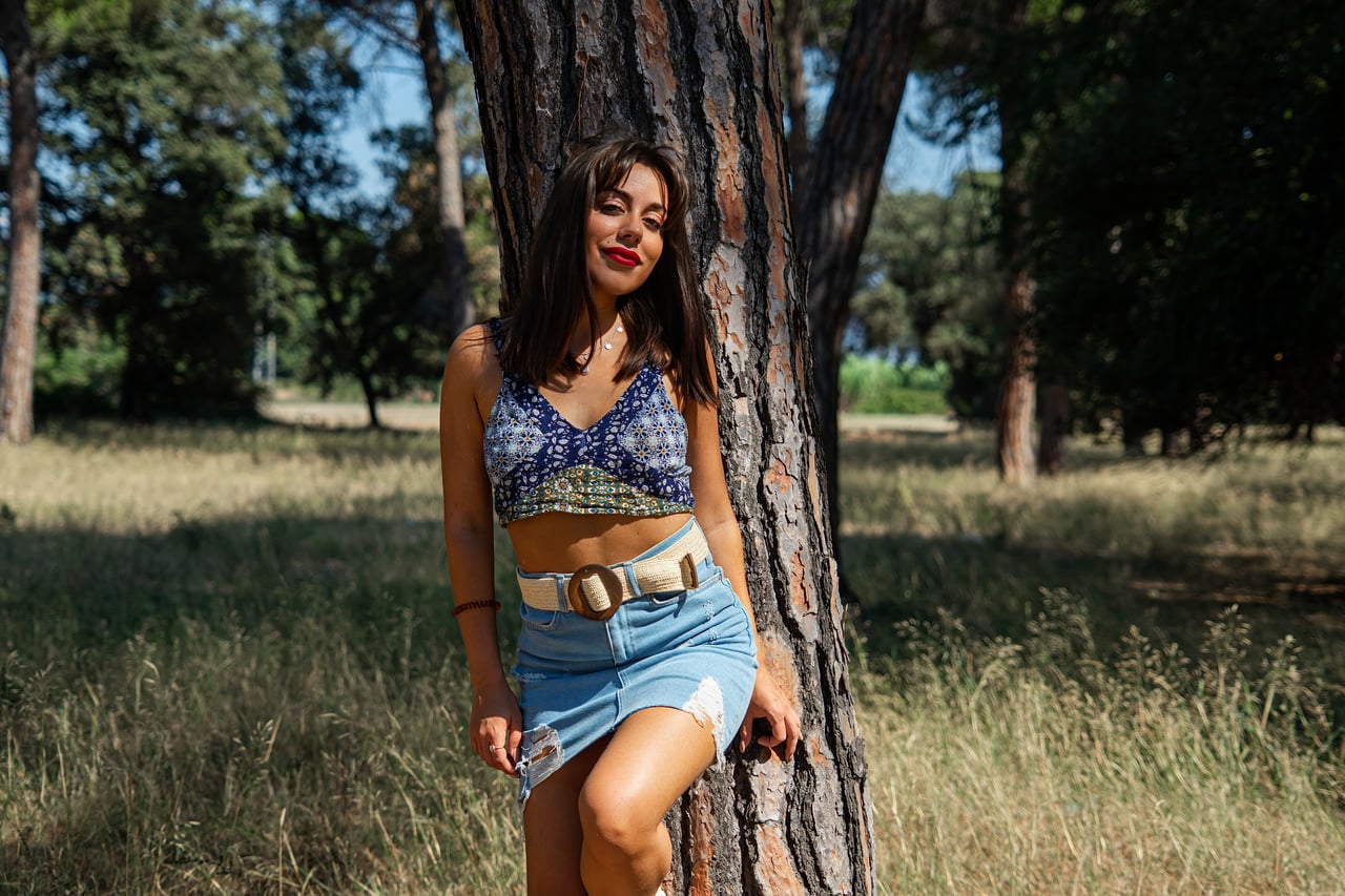Odei8 posa de cos sencer al costat d'un arbre, amb top estampat blau i faldilla texana.