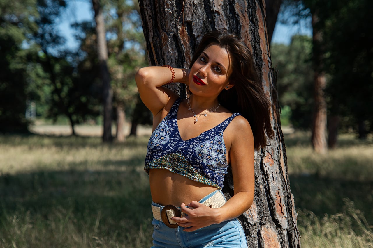 Odei8 posa entre arbres amb top blau estampat, una mà a la cintura i fons verd.