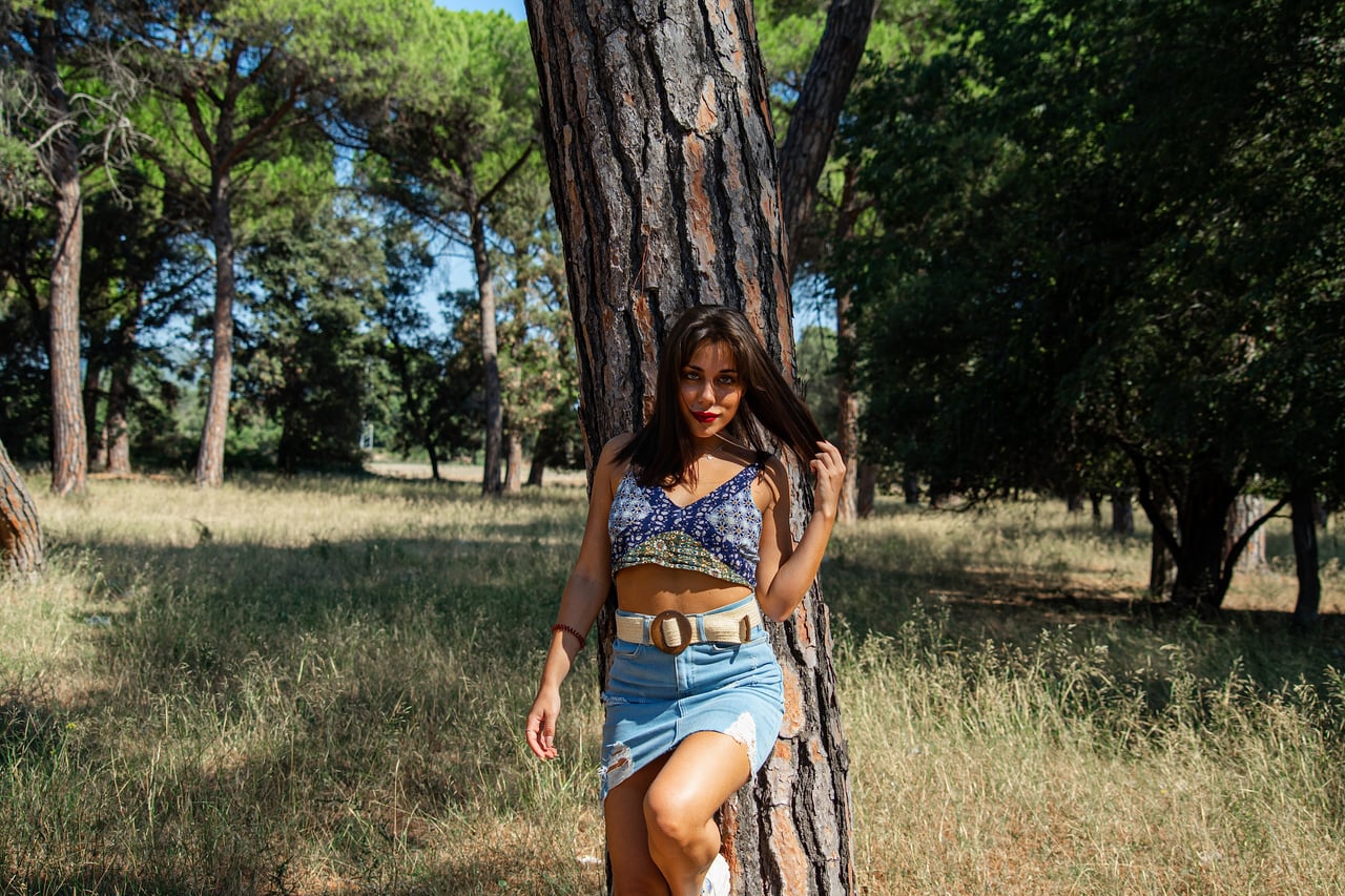 Odei8 posa de cos sencer al costat dels arbres, amb top blau, faldilla texana i mirada a càmera.