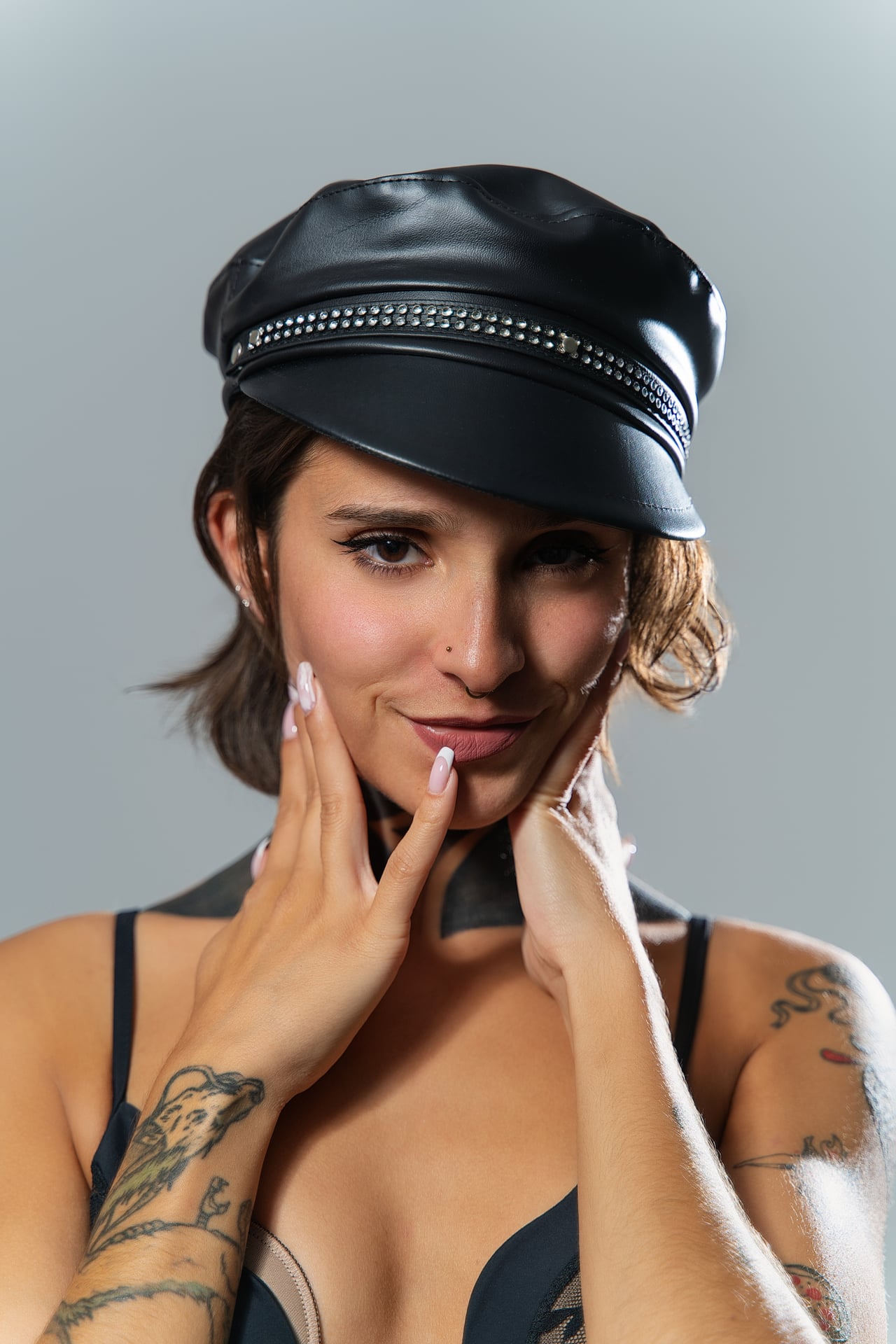 Primer pla de la Rosa amb una gorra de plat de cuir negre i les mans emmarcant el rostre, somrient lleugerament a la càmera, amb bralet de puntes negre i tatuatges als braços, sobre un fons gris llis.