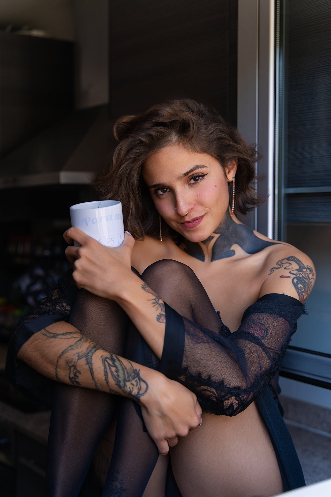 La Rosa posa de mig cos abraçant les cames amb bata de puntes negra transparent i mitges, sostenint una tassa, amb els braços tatuats en primer pla i llum natural lateral.