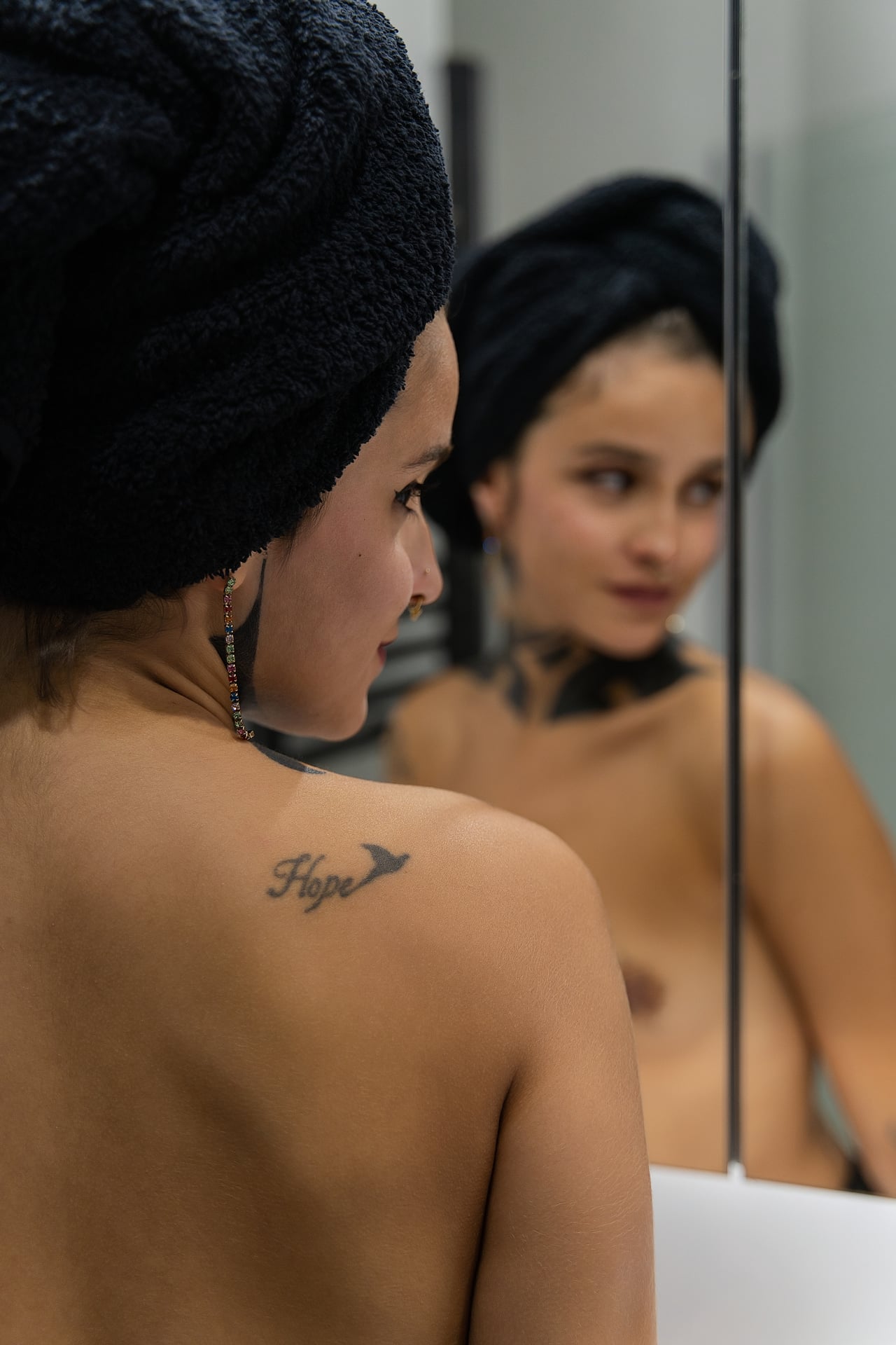 Primer pla de Rosa de perfil amb una tovallola negra al cap i un tatuatge que diu 'Hope' a l'omòplat, d'esquena davant del mirall del bany on es reflecteix el seu rostre i el seu tors descobert.