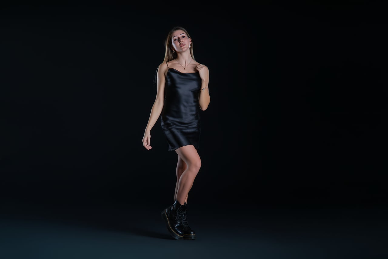 La Sara Cabodevilla avança en moviment subjectant un tirant del vestit, enquadrament de cos sencer, vestit lenceria curt de setí negre i botes militars, sobre fons fosc d'estudi amb llum de flaix low-key.