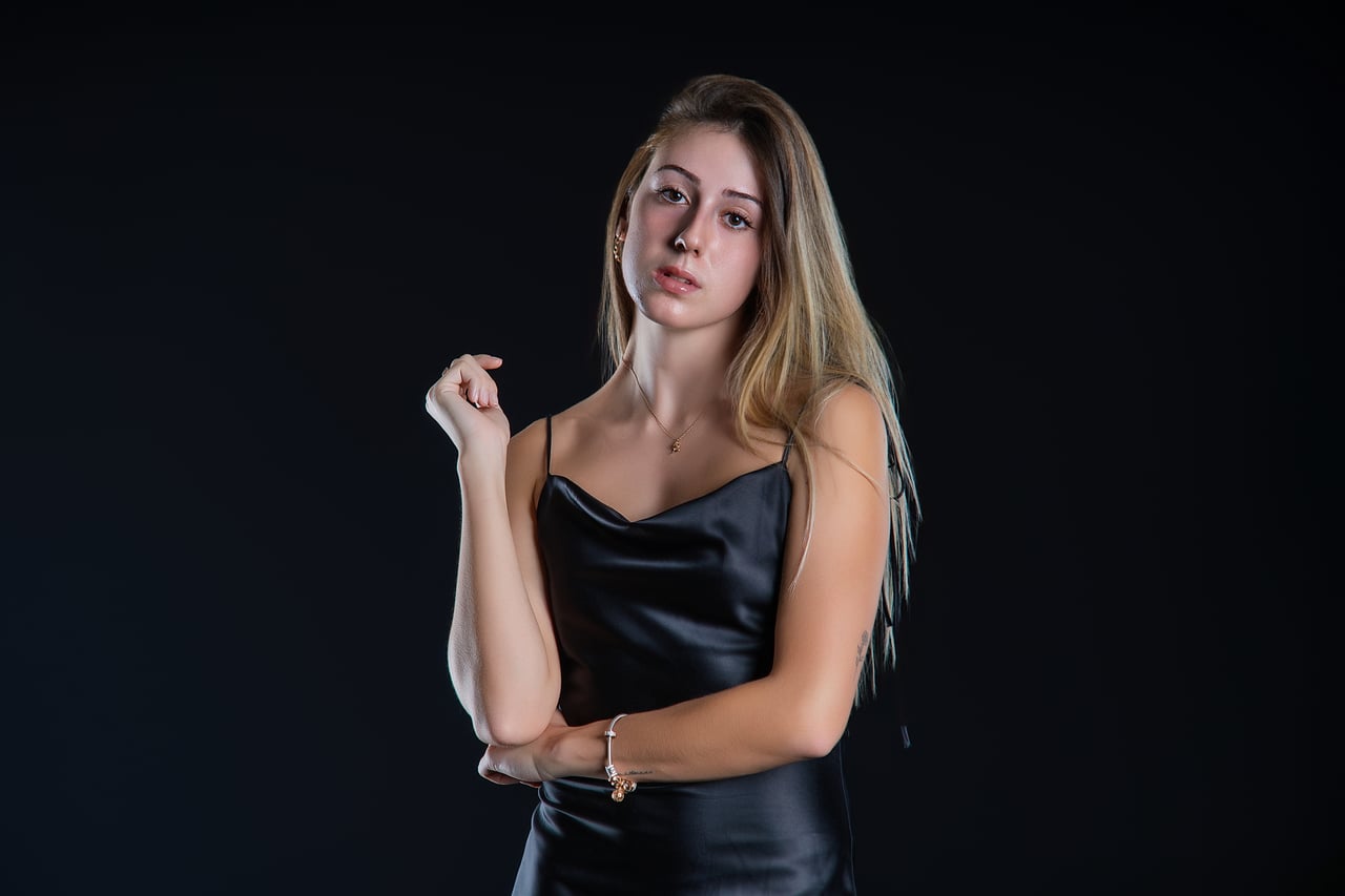 La Sara Cabodevilla mira a la càmera amb un braç flexionat davant del pit, enquadrament de mig cos, vestit lenceria de setí negre, sobre fons fosc d'estudi amb llum de flaix low-key.