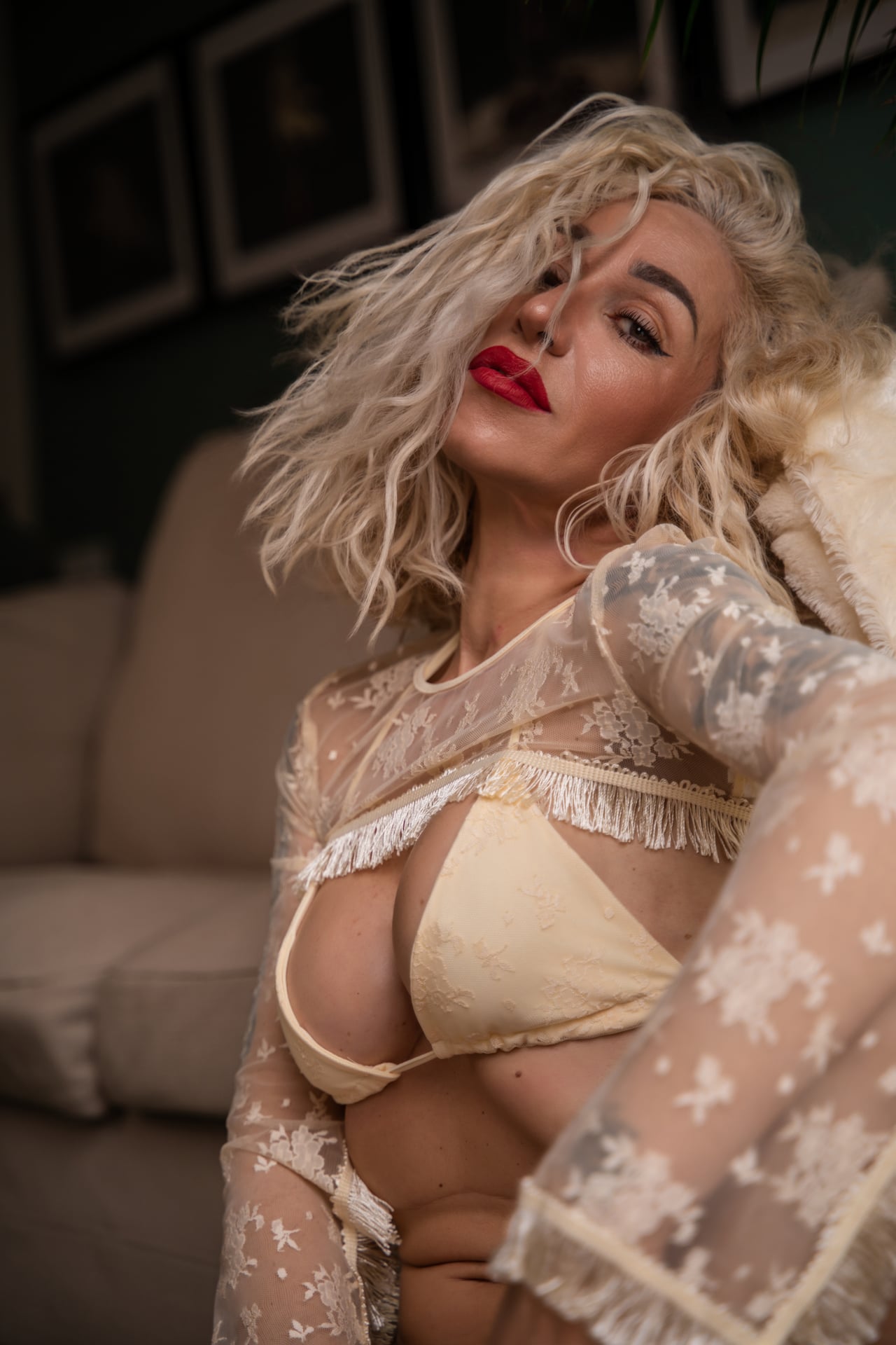 Theresa Rei de mig cos amb la cabellera rossa moguda i una mà al clatell, vestida amb un bralette de puntes crema de copes opaques que cobreix el pit i bata de puntes transparent; pla mitjà amb càlida llum natural i quadres difuminats a la paret del pis.