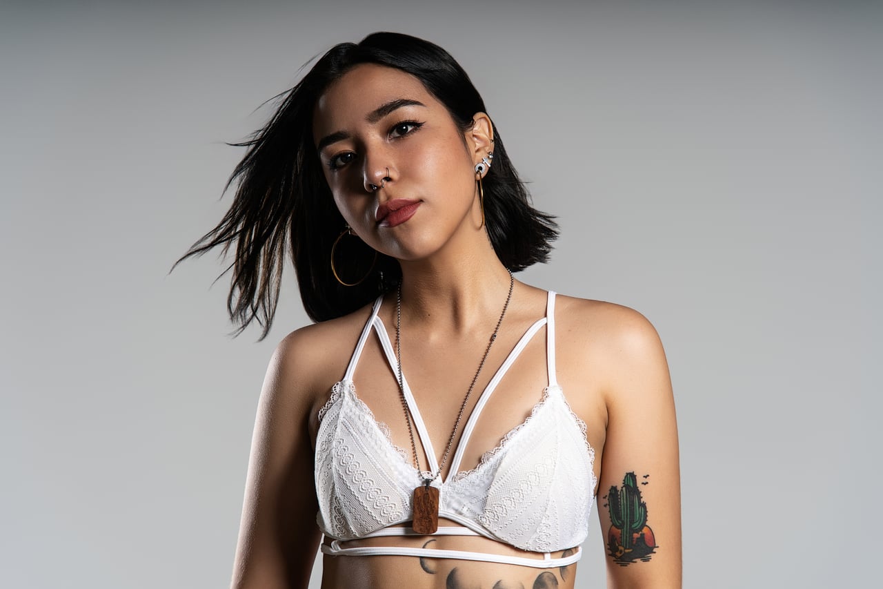 Dona de cabell fosc i melena curta posant de mig cos amb mirada directa, vestida amb un sostenidor de puntes blanc amb tires creuades i penjoll de fusta, tatuatge de cactus al braç, sobre fons gris seamless d'estudi amb llum de flaix controlada.