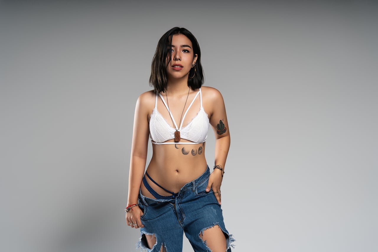 Dona de cabell fosc i melena curta dreta amb mirada directa, vestida amb un sostenidor de puntes blanc amb tires creuades i penjoll de fusta i texans esquinçats abaixats fins als malucs que deixen veure la roba interior, tatuatges a l'abdomen i al braç, sobre fons gris seamless d'estudi amb llum de flaix controlada.
