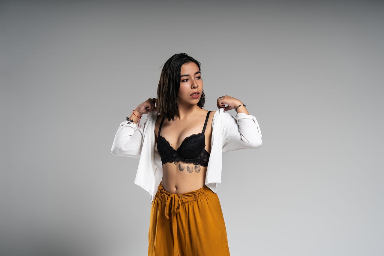 Dona de cabell curt fosc dempeus retirant-se una camisa blanca oberta de les espatlles, deixant veure un sostenidor de blonda negra i un pantaló ample color mostassa, davant d'un fons gris llis amb llum de flaix d'estudi.