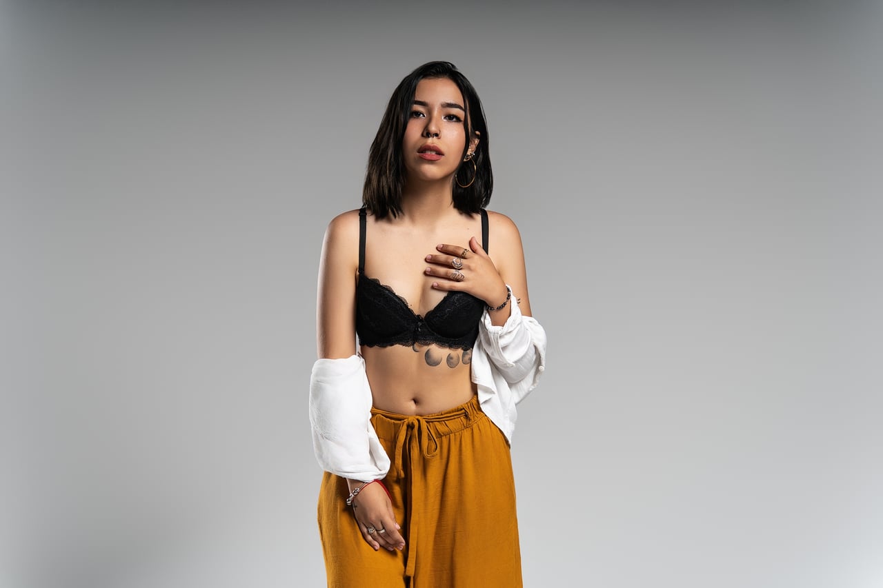 Dona de cabell curt fosc dempeus amb la camisa blanca caiguda sobre els braços i una mà recolzada al pit, lluint un sostenidor de blonda negra i un pantaló ample color mostassa, davant d'un fons gris llis amb llum de flaix d'estudi.