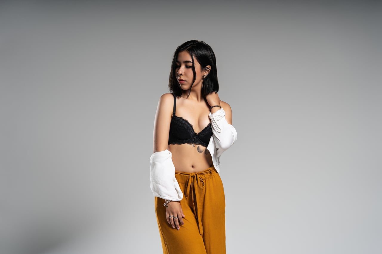 Dona de cabell curt fosc dempeus amb la mirada baixa i la camisa blanca lliscada cap als braços, lluint un sostenidor de blonda negra i un pantaló ample color mostassa, davant d'un fons gris llis amb llum de flaix d'estudi.