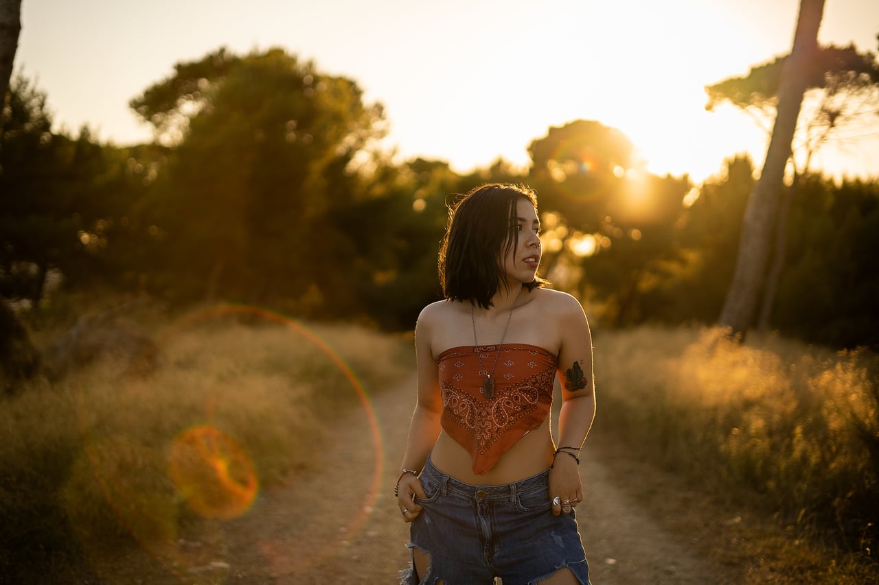 Dona de cabell curt fosc amb top tipus mocador estampat nuat al pit i texans esquinçats posa dempeus en un camí de terra entre vegetació, en enquadrament de cos sencer amb reflexos de sol i llum càlida de capvespre a contrallum.