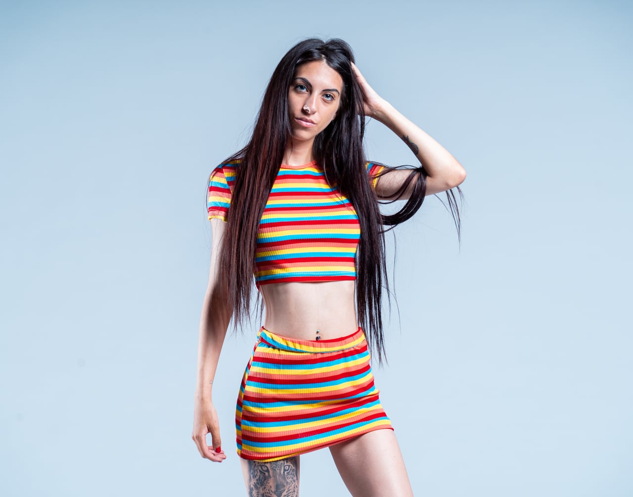 Dona dempeus amb top curt i minifaldilla de ratlles multicolor, mà al cabell, fons d'estudi blau grisenc.